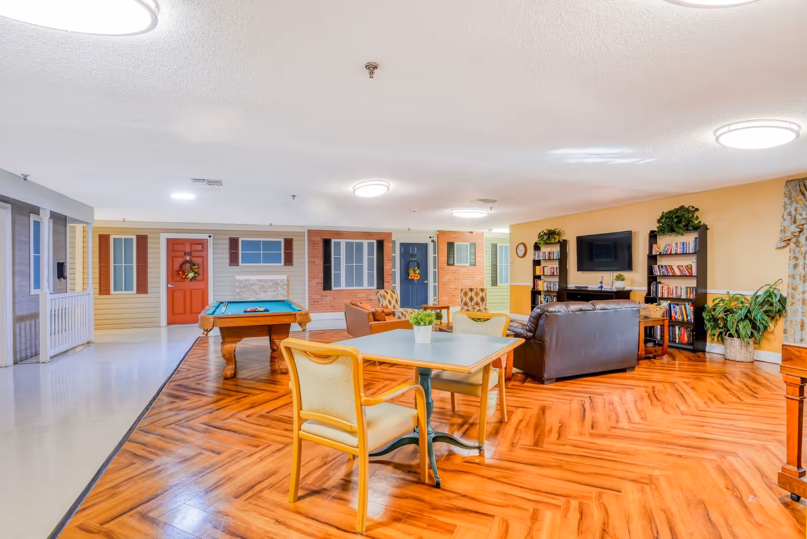 A spacious common area in an assisted living facility featuring a pool table, a seating area with leather couches and armchairs, bookshelves filled with books, a wall-mounted TV, and a table with chairs. The room has wood-patterned flooring and walls decorated to resemble house fronts with doors and windows.