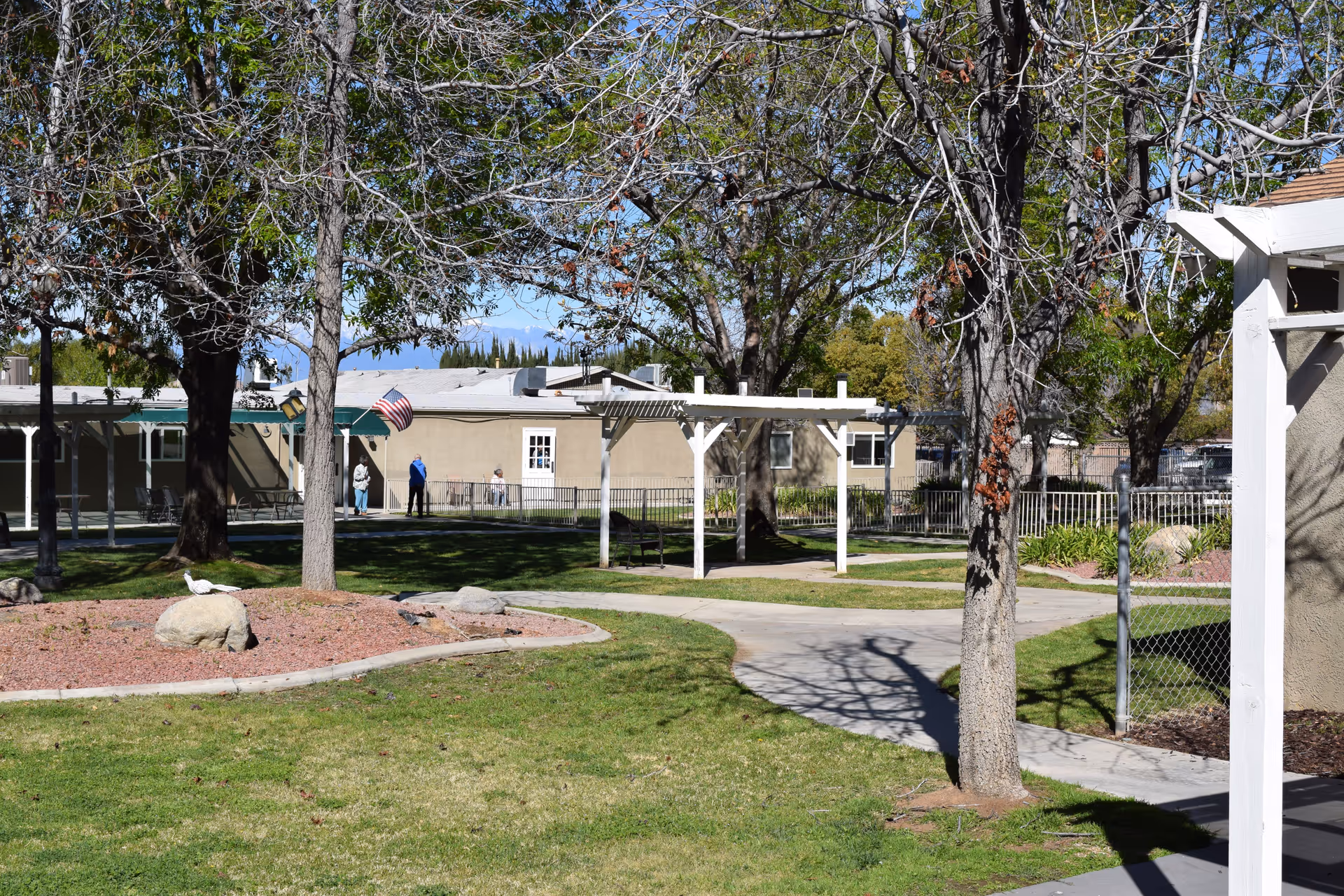 Outdoor garden area at Rose Garden Residential Care with green grass, leafless trees, a paved walkway, a white pergola, and a beige building in the background. There are a few people standing near the building and an American flag is visible.