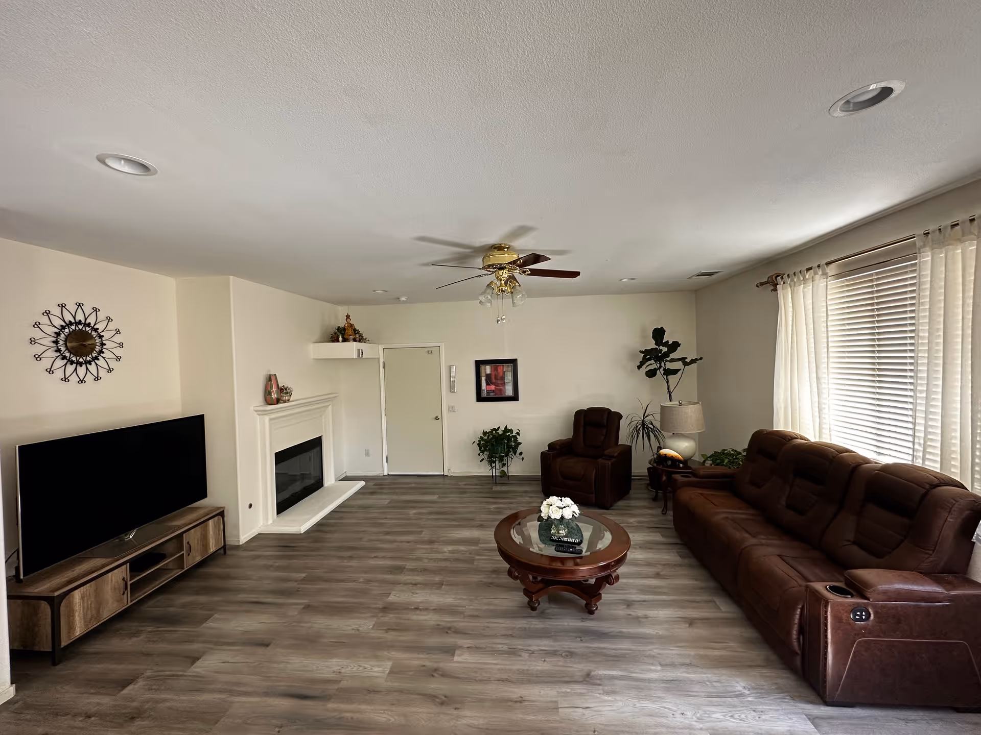 A spacious living room with a brown leather sofa and matching armchair, a round wooden coffee table with a flower vase, a flat-screen TV on a wooden stand, a white fireplace, a ceiling fan with lights, and large windows with white curtains allowing natural light in.