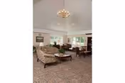 A spacious senior living common room with patterned carpet, upholstered sofas and chairs, a coffee table, a chandelier, and a dining table by windows.