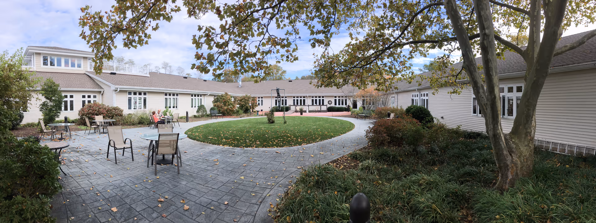 Outdoor courtyard area at The Guilford House featuring a circular grassy lawn surrounded by a paved walkway with several tables and chairs. The courtyard is enclosed by a single-story building with multiple windows and trees providing shade.