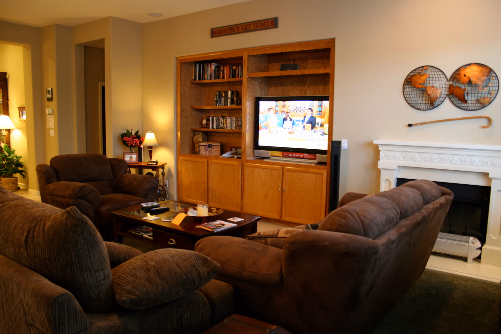 Cozy living room with brown upholstered sofas and armchairs around a coffee table facing a TV in a wooden built-in cabinet next to a white fireplace.