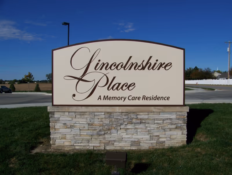 A stone-based outdoor sign on grass reading "Lincolnshire Place - A Memory Care Residence" against a clear blue sky.