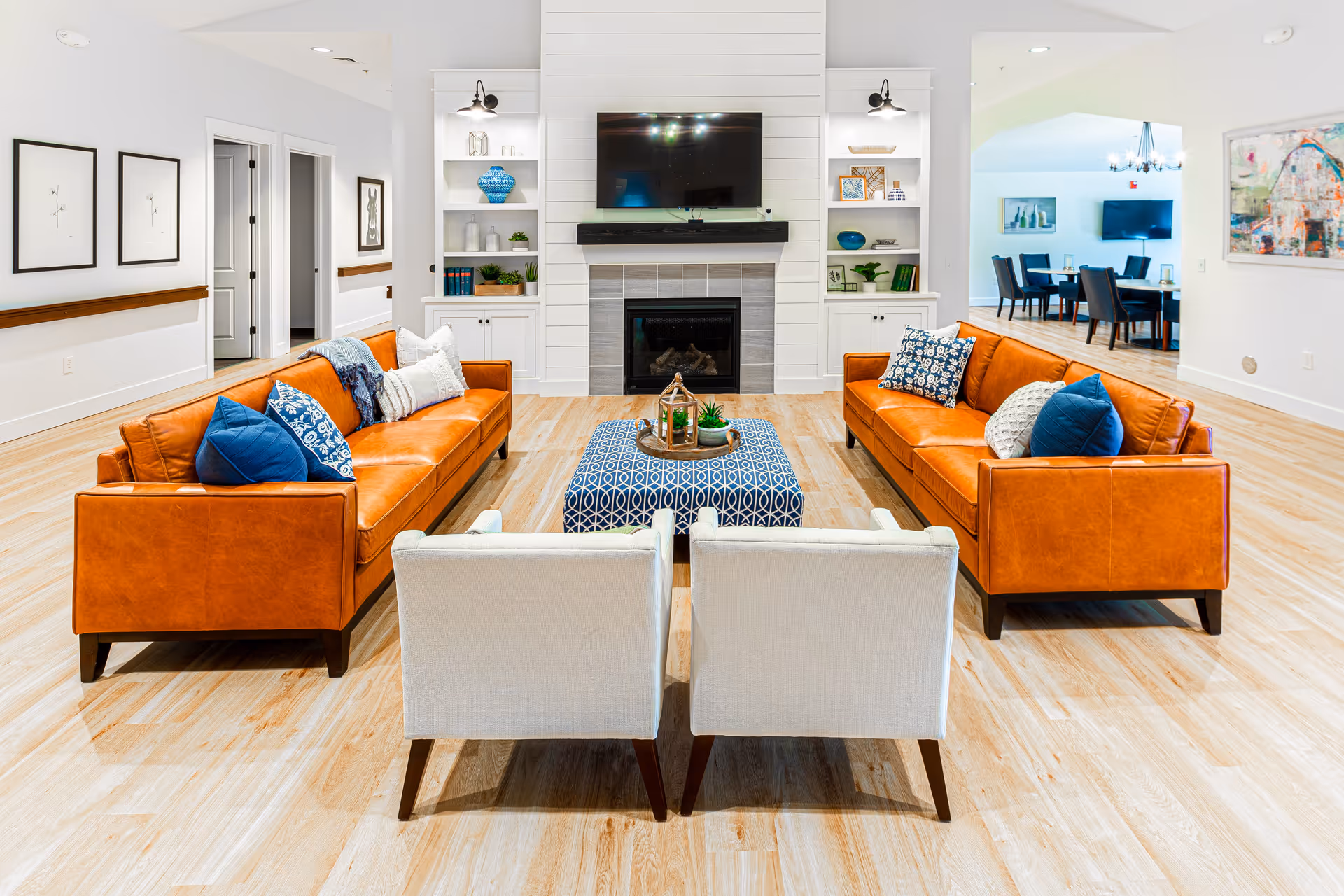 A bright and spacious living room with two orange leather sofas facing each other, two white armchairs, and a blue patterned ottoman in the center. The room features a fireplace with a mounted flat-screen TV above it, built-in white shelves with decorative items, and light wood flooring. In the background, there is a dining area with a table and chairs, and artwork on the walls.