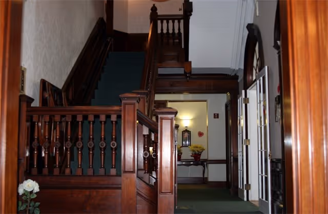 Interior foyer with an ornate dark-wood staircase and banister, green carpet, and a hallway table with flowers.