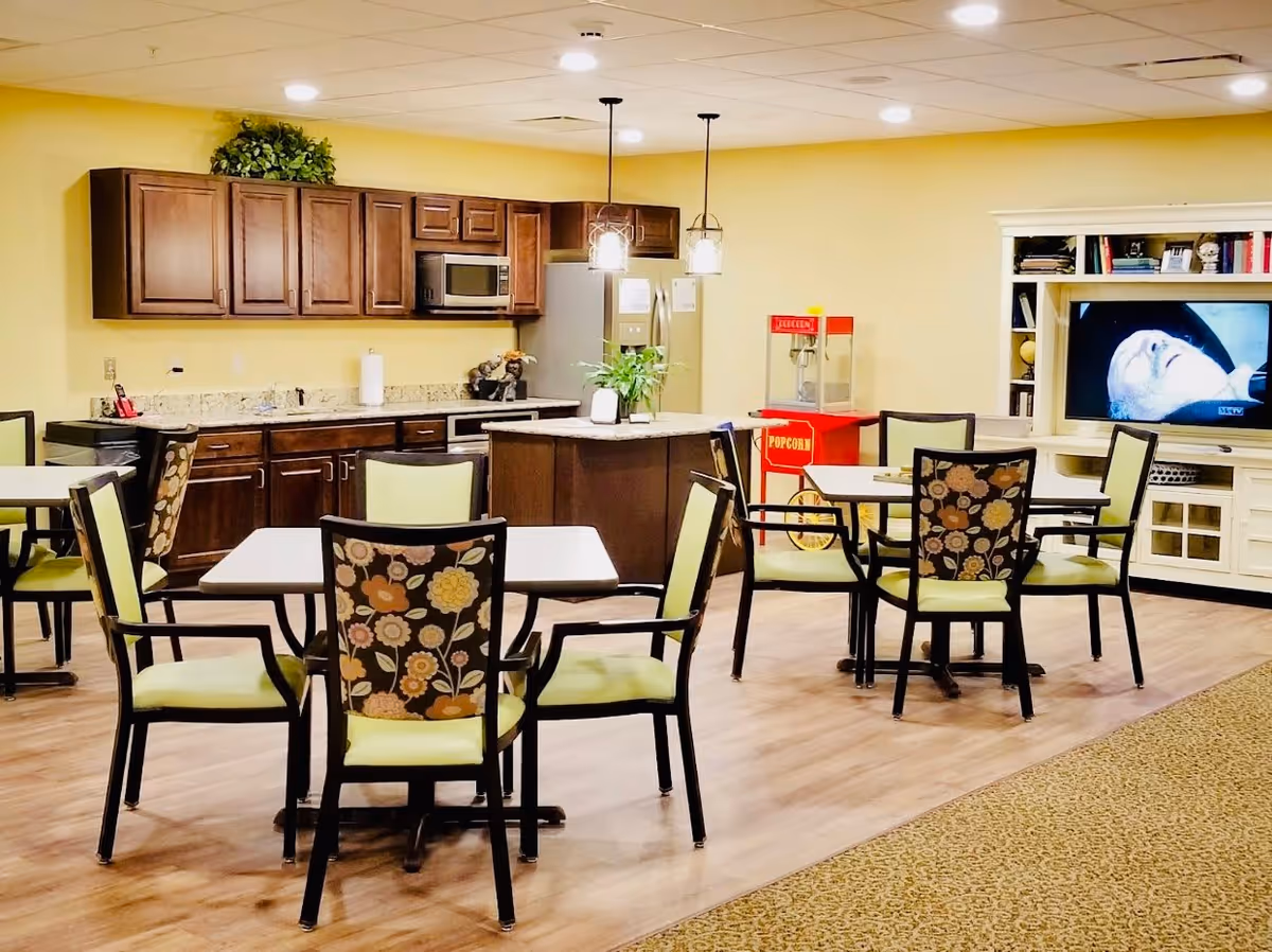 A communal dining and kitchen area in a senior living facility with several tables and chairs, a kitchen with wooden cabinets, a microwave, and a refrigerator. There is a popcorn machine and a television showing a close-up of a person's face. The room has warm yellow walls and wood flooring.