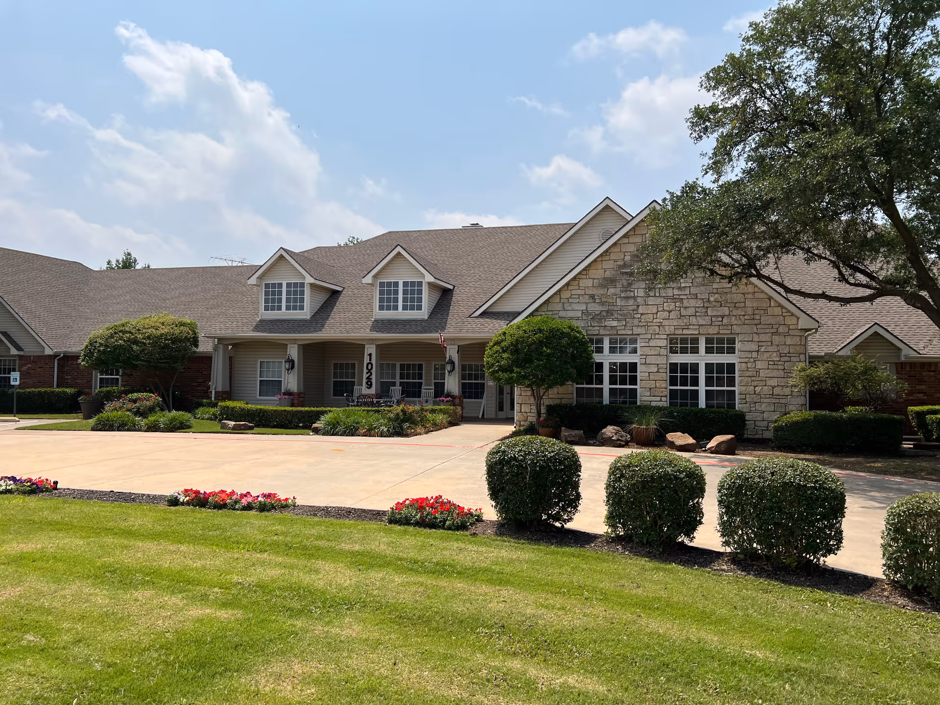 Front exterior view of a single-story building with a stone and brick facade, multiple windows, and a covered porch area. The building is surrounded by neatly trimmed bushes, a green lawn, and flower beds under a partly cloudy sky.