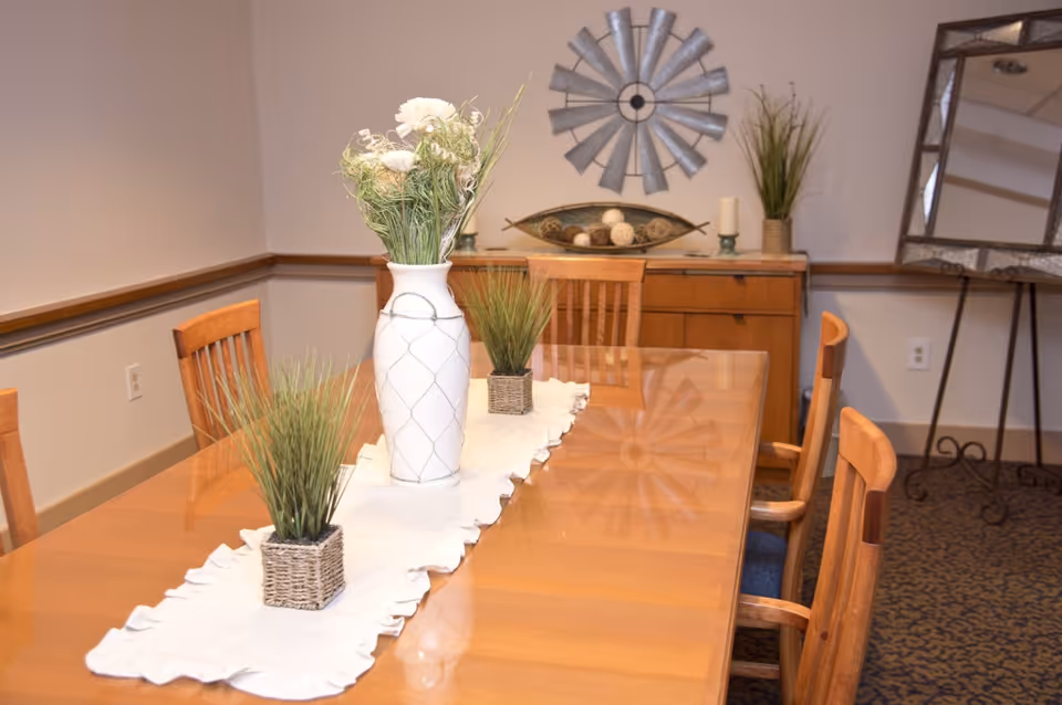 A dining room with a wooden table and six wooden chairs. The table has a white runner with a white vase holding flowers and two small potted plants. In the background, there is a wooden sideboard with decorative items including a metal windmill wall decoration, candles, and a plant. A large framed mirror is on the right side of the room.