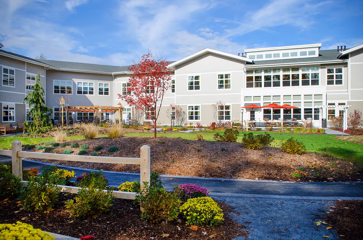 Exterior view of a senior living facility building with multiple windows, a landscaped garden with colorful flowers, shrubs, a small tree with red leaves, a wooden fence, and a paved walkway. There are outdoor seating areas with red umbrellas near the building.