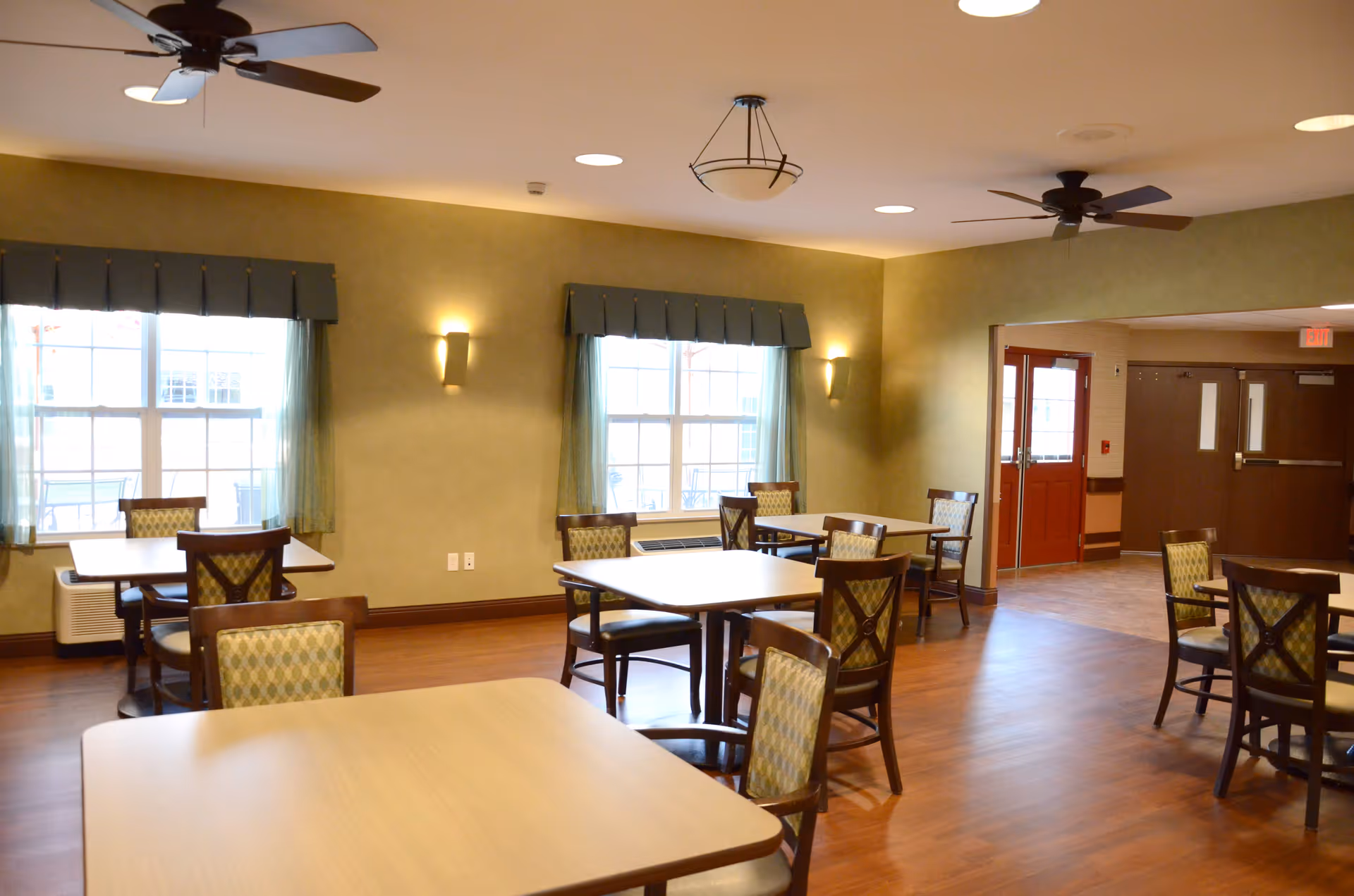 Communal dining room with several wooden tables and upholstered chairs, ceiling fans, windows with valances, and wood-look flooring.