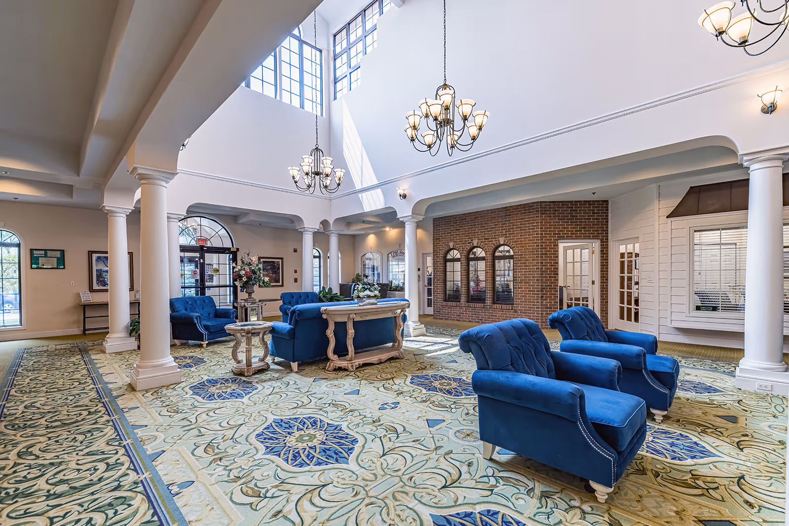 Spacious senior living lobby with blue upholstered chairs, columns, chandeliers, and patterned carpet under a high skylight.