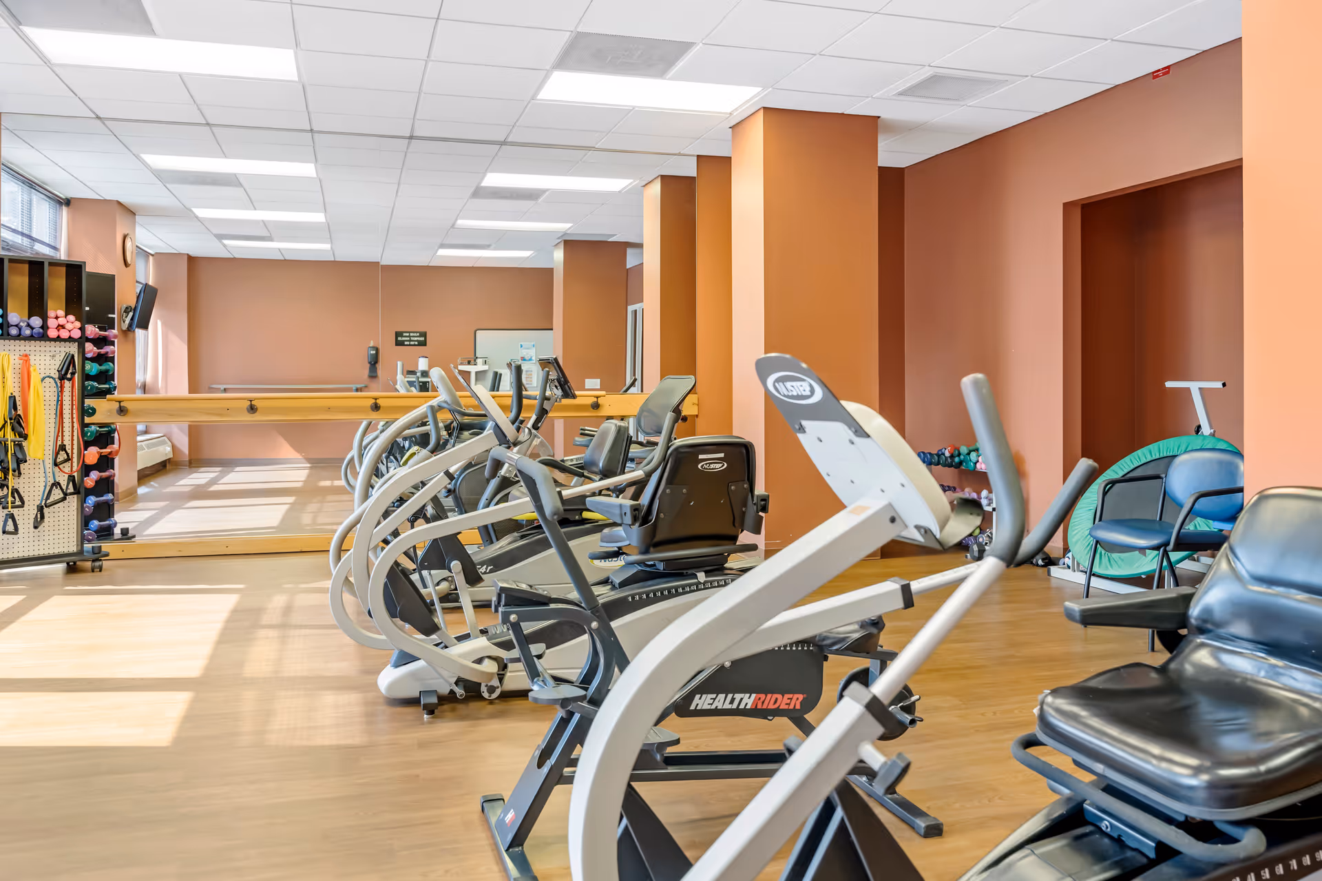 Exercise room with several stationary bikes and other fitness equipment, a rack with colorful dumbbells and resistance bands, large mirrors on the wall, and orange-colored walls with wooden flooring.