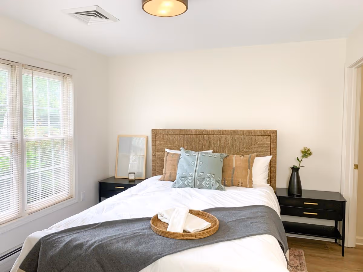 A bright bedroom with a large bed featuring a woven headboard, white bedding, and decorative pillows in blue and brown tones. A gray throw blanket is draped across the foot of the bed with a round wicker tray holding folded white towels. On either side of the bed are black nightstands; the left one has a framed mirror and a small clock, and the right one has a black vase with a green plant. A large window with white blinds allows natural light to fill the room.