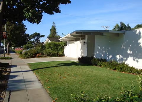Single-story white mid-century style house with a front lawn, driveway and sidewalk lined with trees under a clear blue sky.
