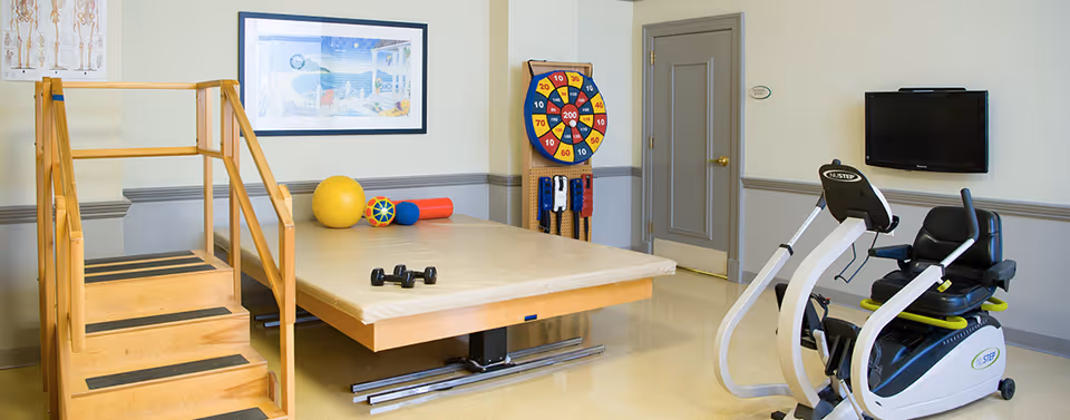 Therapy/exercise room with a raised treatment table and steps, exercise bike, therapy balls, and a wall-mounted TV.