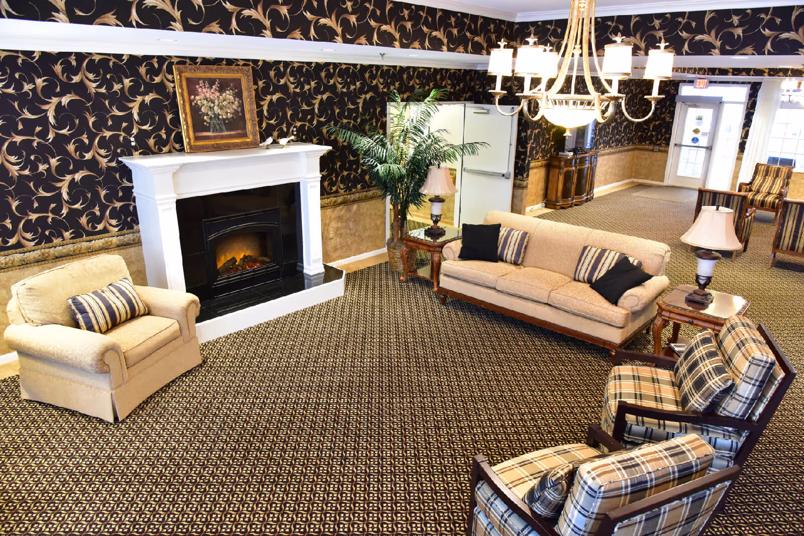 A cozy living room area in an assisted living facility featuring a beige sofa with striped pillows, a matching armchair, two plaid armchairs, a white fireplace with a floral painting above it, a potted plant, side tables with lamps, and a chandelier hanging from the ceiling. The room has patterned carpet and wallpaper with a dark floral design.