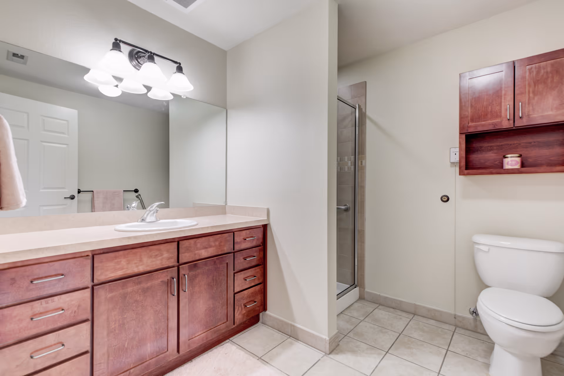 A clean bathroom featuring a large mirror above a wooden vanity with a sink, a toilet, a glass-enclosed shower, and a wooden cabinet mounted on the wall. The floor is tiled and the walls are painted light beige.