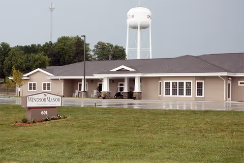 Exterior view of a single-story assisted living facility building with a sign in front that reads Windsor Manor Assisted Living. The building has a covered entrance and multiple windows. A water tower is visible in the background behind the building. The surrounding area includes a grassy lawn and some trees.