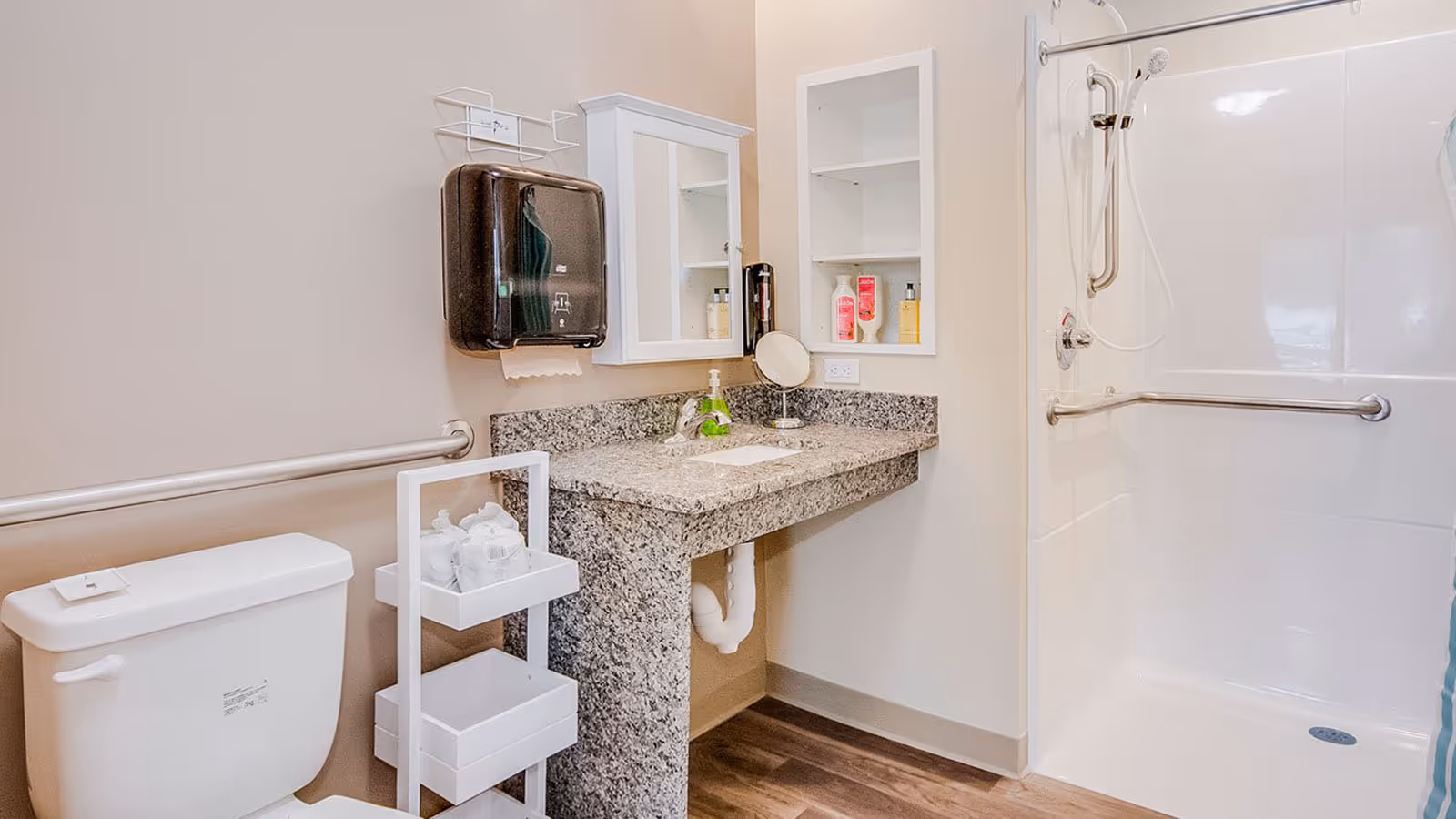 A clean and accessible bathroom featuring a white toilet, a granite countertop with a built-in sink, a wall-mounted paper towel dispenser, a small mirror cabinet, and an open shelving unit with toiletries. The shower area has a handheld showerhead, grab bars, and a white shower curtain with blue accents. The floor is wood-style laminate.