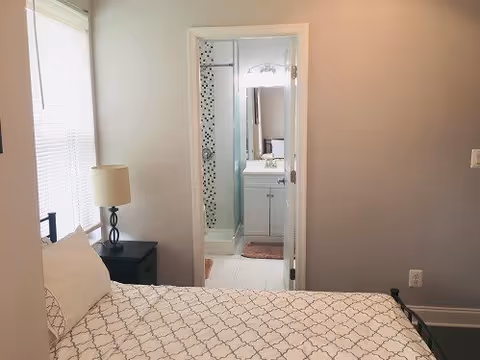A bedroom with a bed covered in a white and gray patterned bedspread, a black nightstand with a lamp, and a window with blinds. Through an open door, a bathroom is visible with a glass shower door, a white vanity with a sink, and a mirror above it.