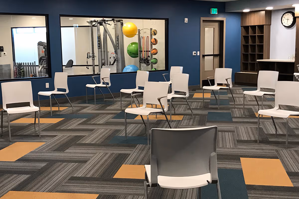 Spacious interior activity room with white chairs arranged in spaced rows on patterned carpet and a view into an adjacent fitness area with exercise balls.