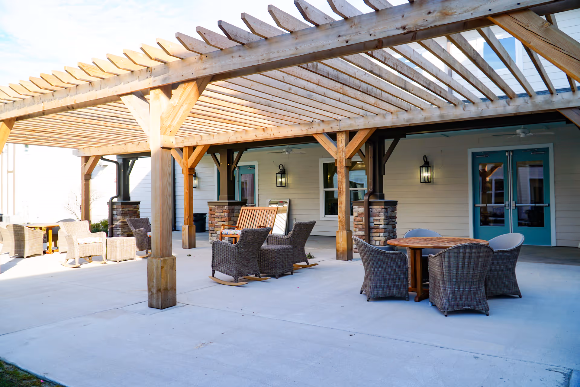 Outdoor patio area at Seagrass Village of Gulf Shores featuring a wooden pergola, wicker chairs, a wooden rocking chair, and a round wooden table with four wicker chairs. The patio is adjacent to a building with teal double doors and windows.