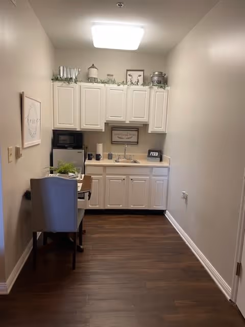 A small kitchenette area with white cabinets, a sink, a microwave, and a mini refrigerator. There is a small table with a gray upholstered chair on the left side. The countertop has a few decorative items and a sign that says 'FEED'. The floor is dark wood, and the walls are painted beige.