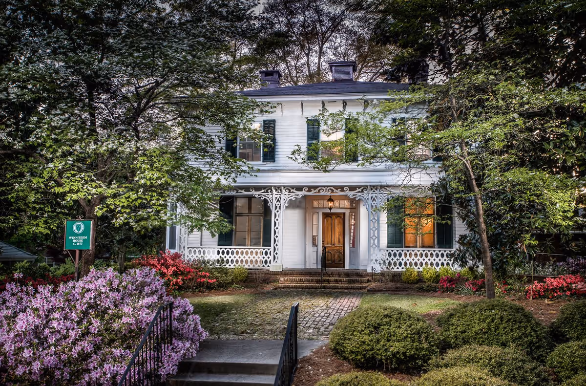 Front exterior view of a two-story white house with green shutters, a wooden front door, and decorative white porch railings. The house is surrounded by lush greenery, flowering bushes, and trees, with a brick pathway leading to the entrance. A green sign near the front reads 'Winn-Teem House C. 1873'.