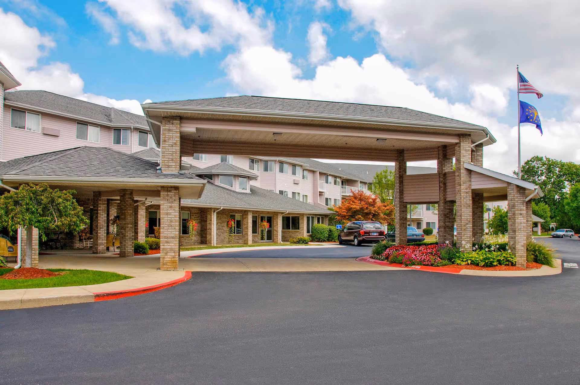 Entrance of Arbor Glen Independent and Assisted Living facility showing a covered drop-off area with brick pillars, a driveway, landscaped flower beds, and two flags on a flagpole against a partly cloudy sky.