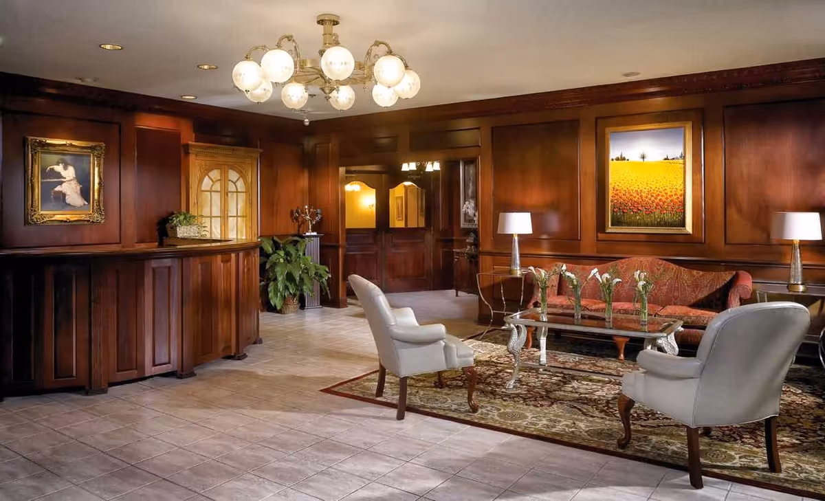 A warmly lit interior lounge area with wooden paneled walls, a chandelier with globe lights, a wooden reception desk, two white armchairs, a red patterned sofa, a glass coffee table with flower vases, two table lamps, framed artwork on the walls, and a decorative rug on the tiled floor.