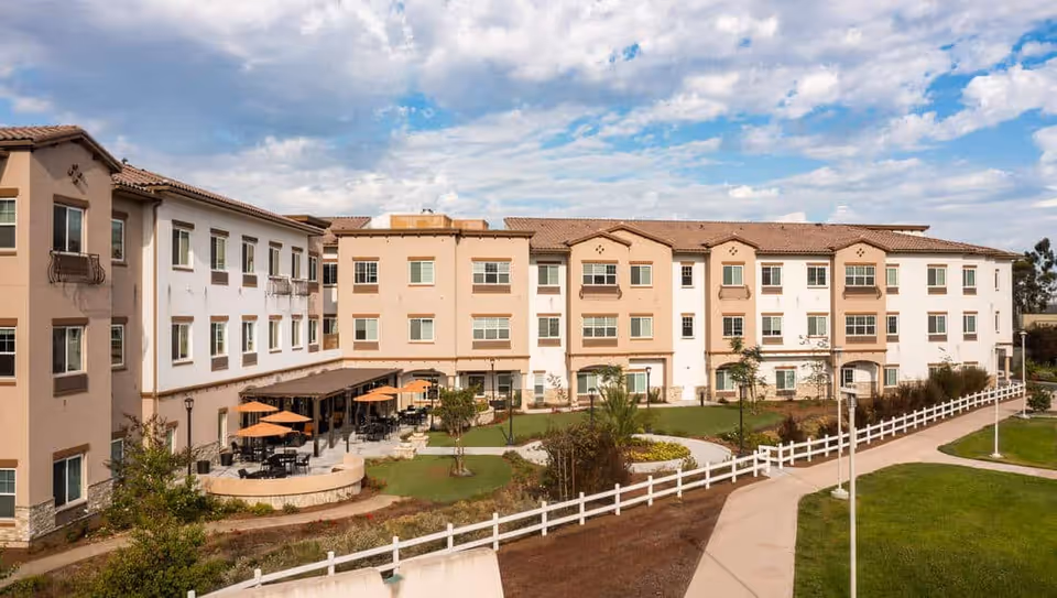 Exterior view of a three-story senior living facility building with beige and white walls, multiple windows, and a tiled roof. The foreground features a landscaped garden area with a curved pathway, green lawns, small trees, and outdoor seating with tables and orange umbrellas. The sky is partly cloudy.