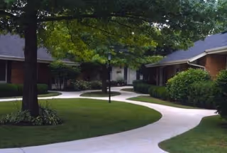 Curved concrete pathways surrounded by well-maintained green lawns and bushes, with single-story brick buildings and large trees providing shade in an outdoor courtyard area.