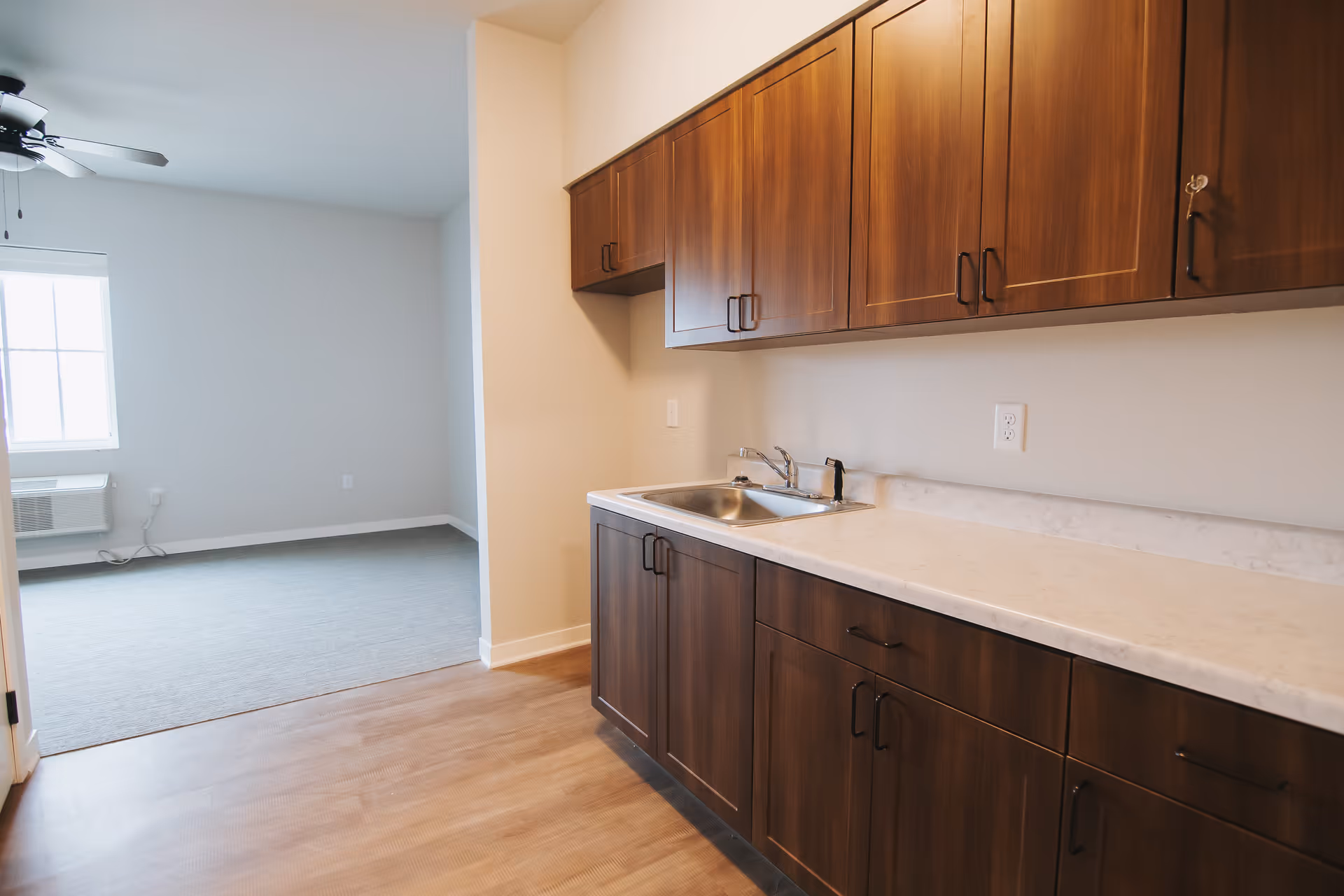 Interior view of a room in a senior living facility showing a kitchenette area with wooden cabinets, a countertop with a sink, and an adjacent carpeted room with a window and ceiling fan.