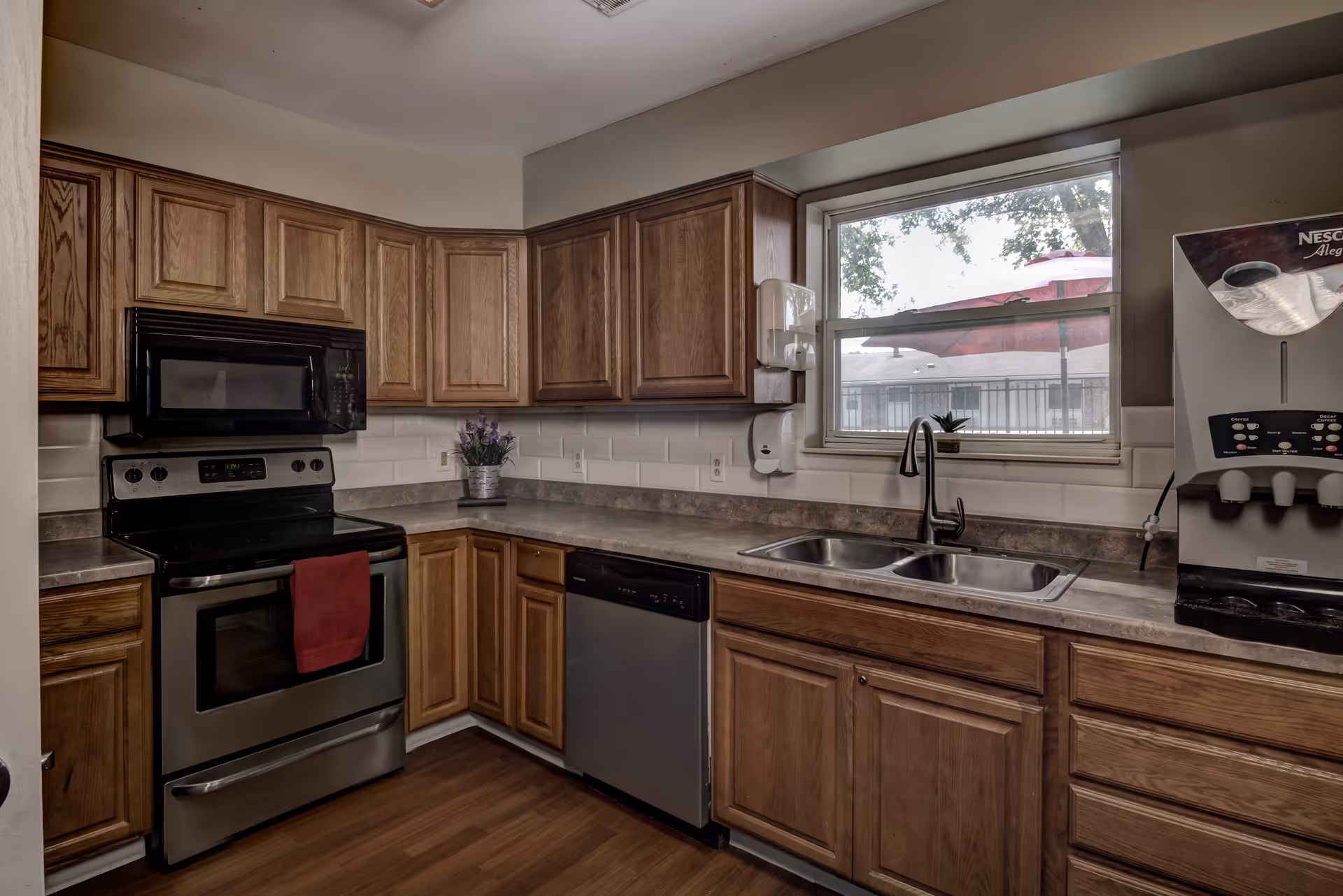 A kitchen with wooden cabinets, a stainless steel oven with a red towel hanging on the handle, a dishwasher, a double sink under a window, and a coffee machine on the right side. There is a small potted plant on the windowsill and a lavender plant on the countertop.