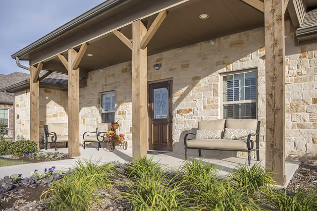 Stone-front porch with wooden posts, cushioned benches, potted flowers, and a glass-paneled front door.