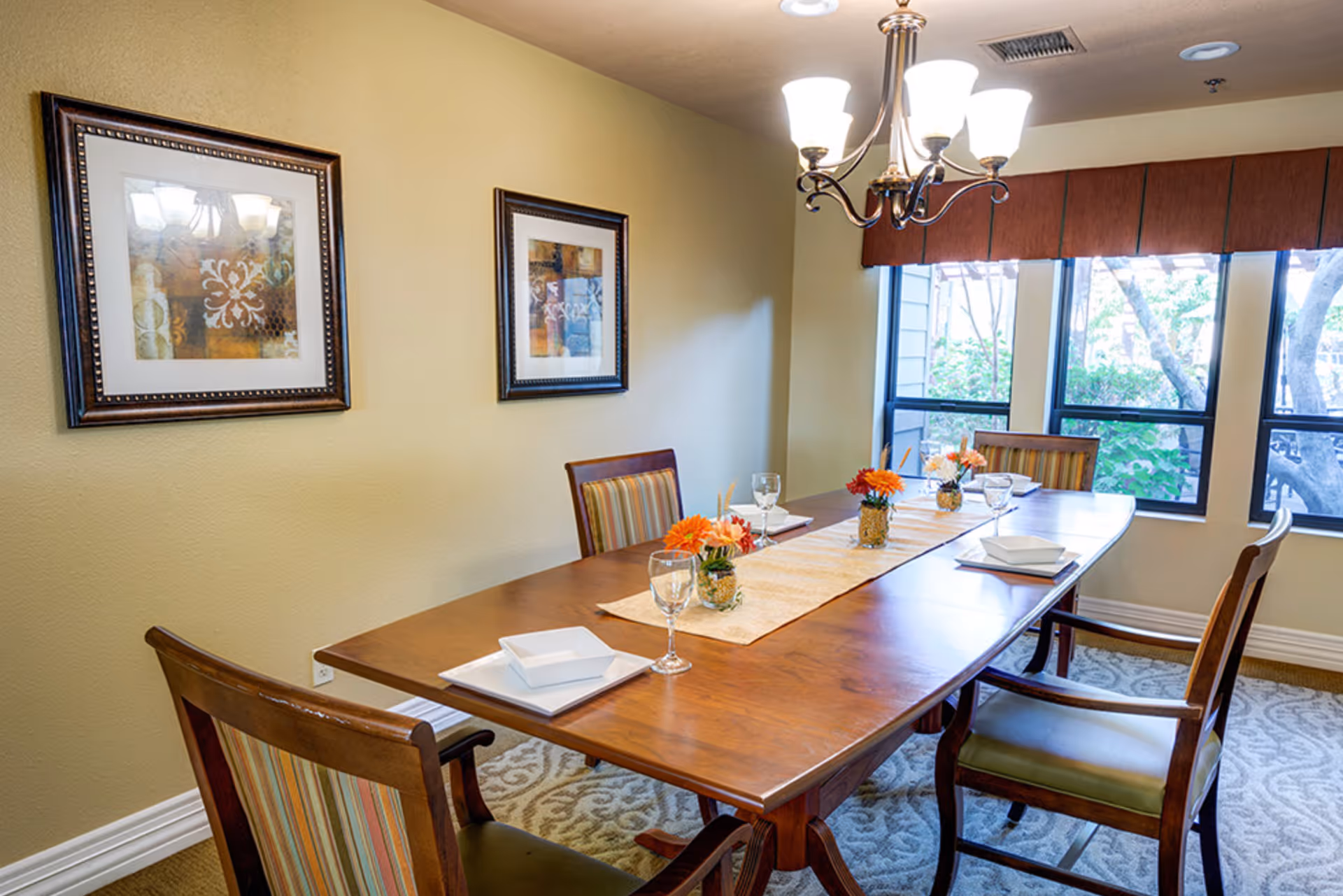 Well-lit dining room with a wooden table set with place settings and small floral arrangements, upholstered chairs, framed artwork, and a chandelier by large windows.