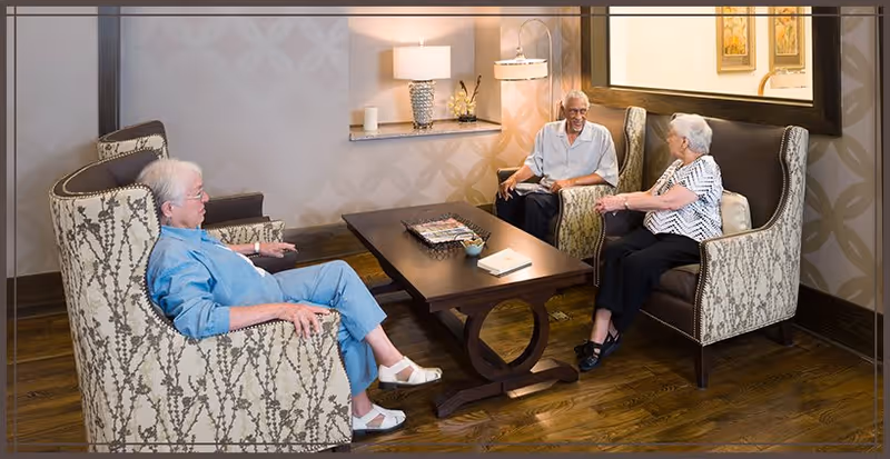 Three elderly women sitting and conversing in a cozy living room area with patterned armchairs, a wooden coffee table, a lamp on a side table, and a large mirror on the wall.