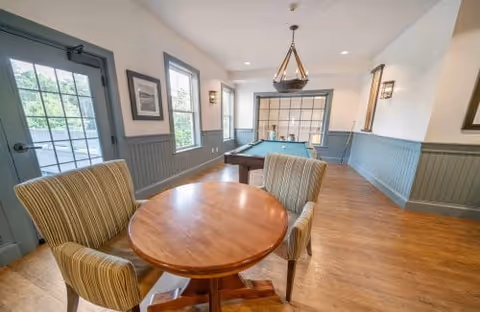 Bright common room with a round table and upholstered chairs in the foreground and a pool table under a chandelier by windows and a glass-paned door.