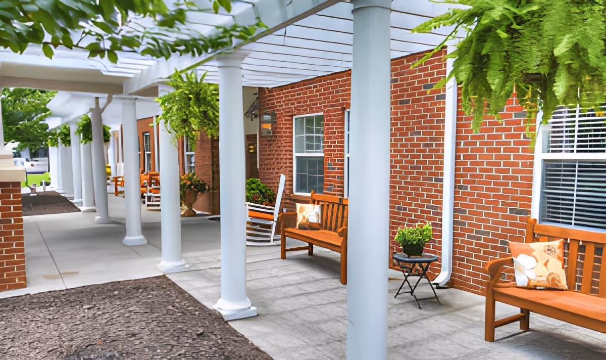 Covered outdoor walkway with white columns and a white pergola roof. There are wooden benches with cushions and small tables with potted plants along a red brick wall. Hanging green ferns are suspended from the pergola. The area is clean and well-maintained, creating a welcoming outdoor seating space.