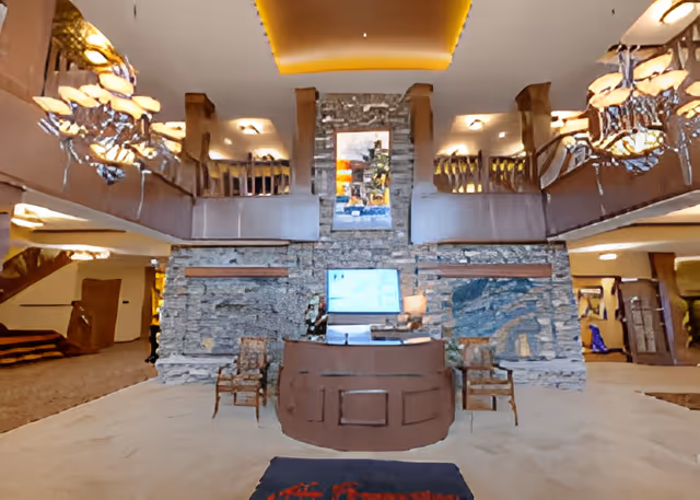 Interior view of a senior living facility lobby with a central stone wall featuring a mounted TV and artwork above it. In front of the wall is a curved reception desk with two wooden chairs on either side. The lobby has a high ceiling with warm lighting, wooden railings on the upper level, and decorative chandeliers hanging from the ceiling.