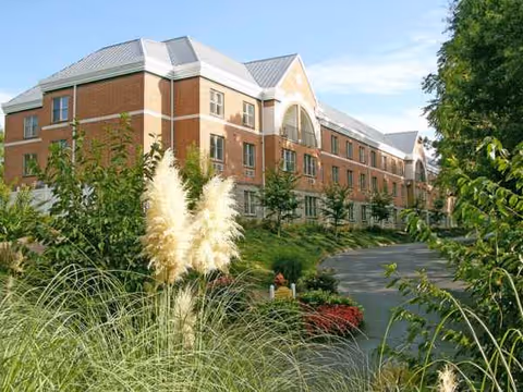 Brick senior living building with arched entrance, surrounded by green landscaping and a winding driveway.