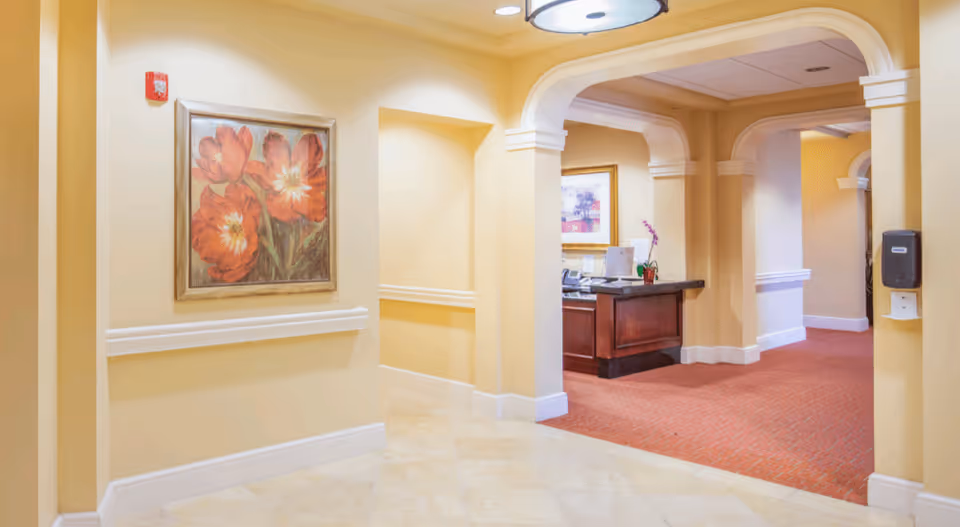 Interior hallway of a senior living facility with beige walls, a framed painting of red flowers on the left wall, arched doorways, and a reception desk with a computer and a small plant in the background. The floor is tiled in the foreground and carpeted in the background.