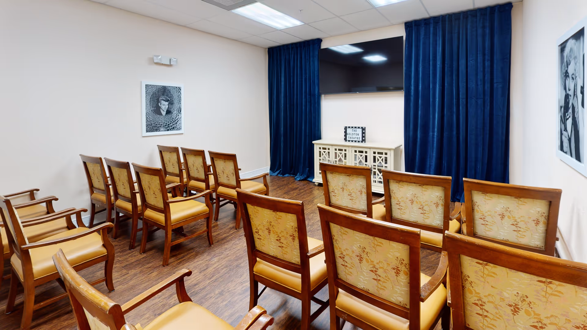 A small theater room with rows of wooden chairs featuring floral upholstery facing a large flat-screen TV mounted on the wall. The TV is framed by dark blue curtains, and there is a white cabinet below it with a sign that reads 'The Goldton Theatre'. The room has wood flooring and light-colored walls with black and white framed photographs.
