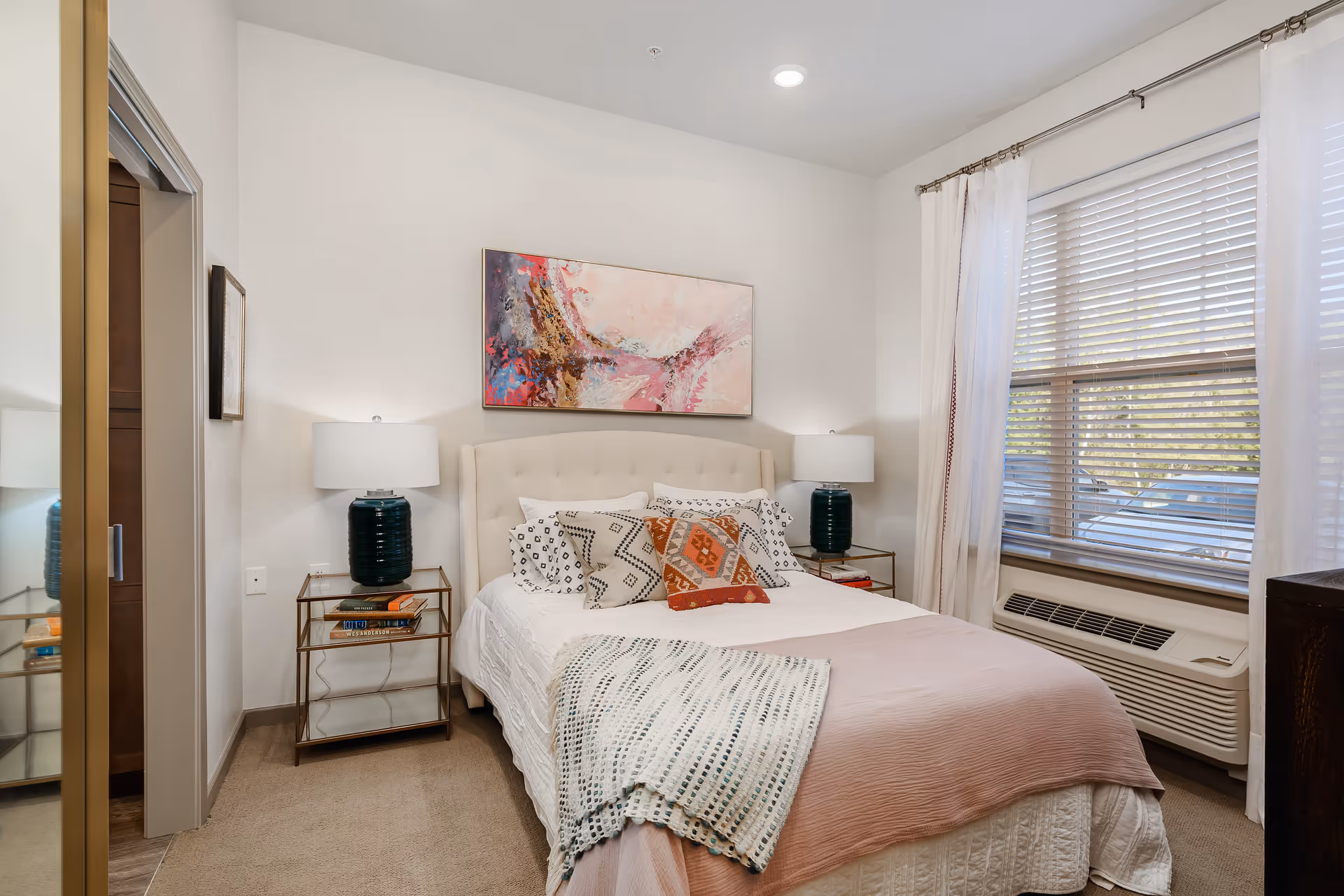 A cozy bedroom with a neatly made bed featuring multiple patterned pillows and a pink throw blanket. There are two matching bedside tables with dark blue lamps on each side of the bed. Above the bed hangs a colorful abstract painting. A large window with white curtains and blinds lets in natural light. The room has beige carpeting and light-colored walls.