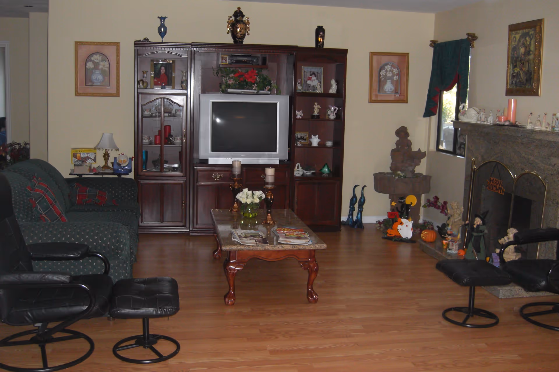 A living room with wooden flooring featuring a dark green couch with red and green pillows, two black leather swivel chairs with matching footrests, a wooden coffee table with a marble top holding candles and a flower vase, and a large wooden entertainment center with a television and decorative items. The room also has a stone fireplace decorated with Halloween-themed figurines and a window with green curtains.