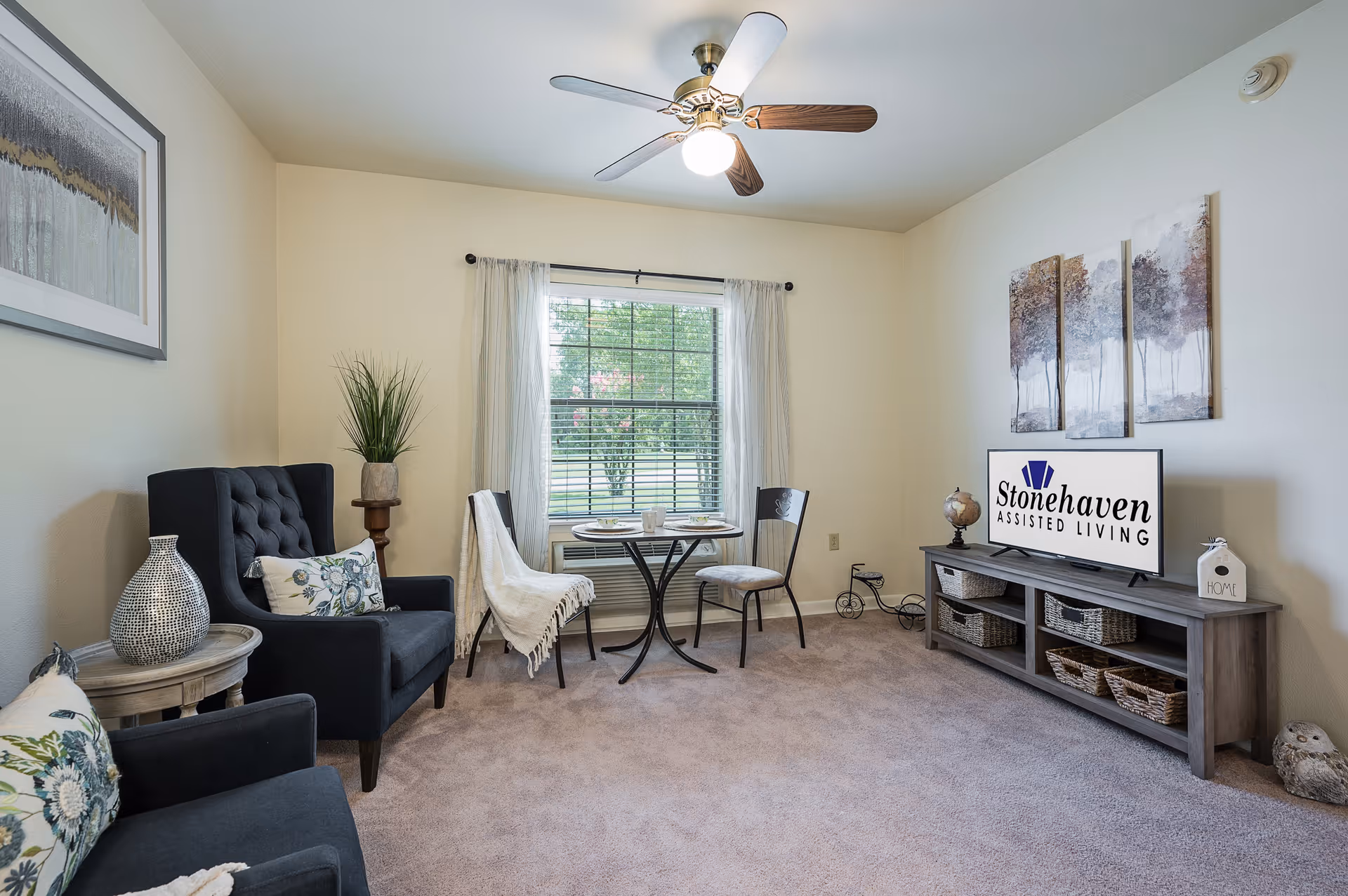Cozy assisted-living living room with two armchairs, a small round table by a window, and a TV on a media console displaying the facility name.