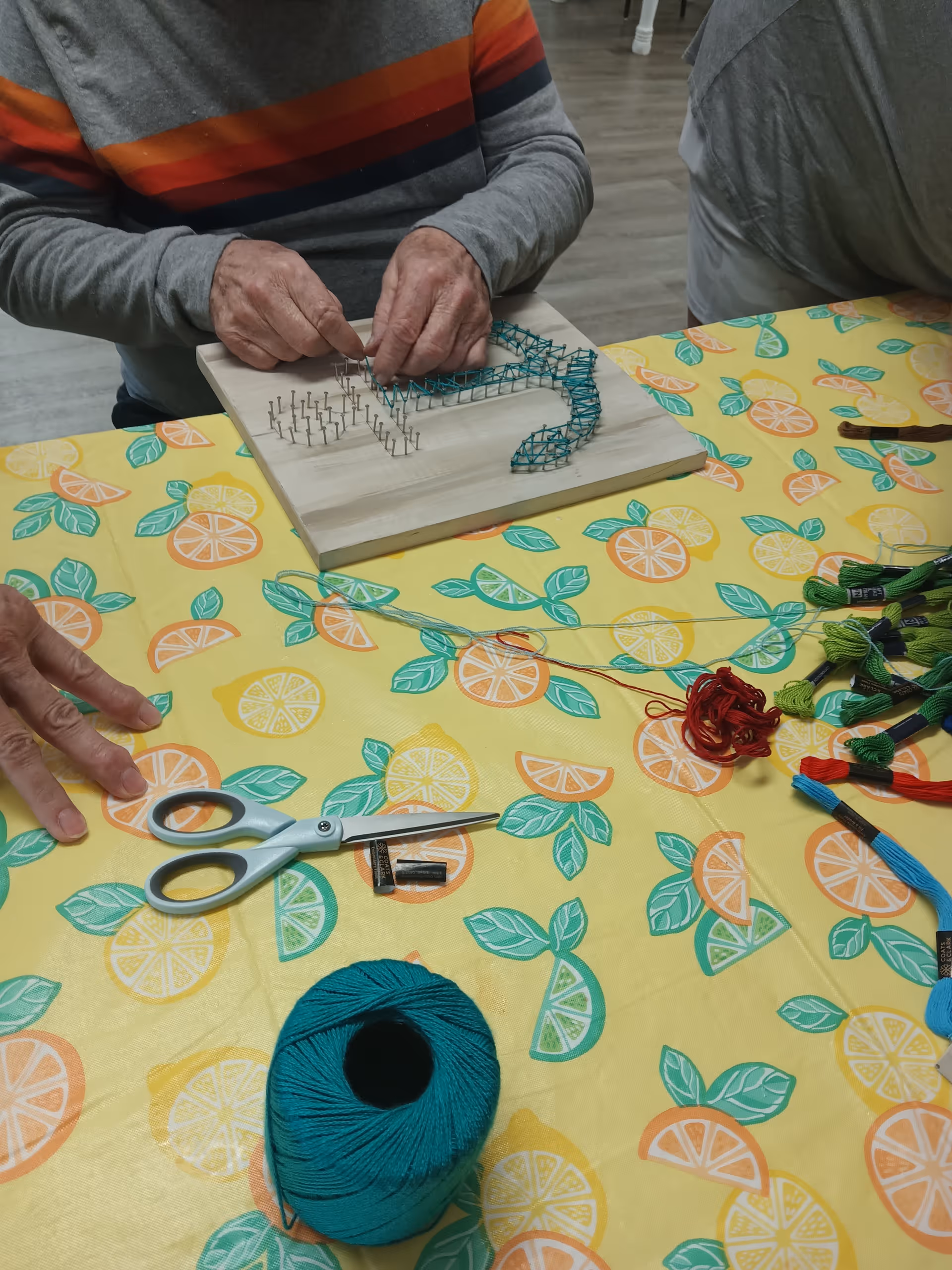 Two people sitting at a table covered with a yellow tablecloth decorated with citrus fruit patterns. One person is working on a string art project using nails and blue thread on a wooden board. Various colored threads, a pair of scissors, and a spool of teal yarn are also on the table.