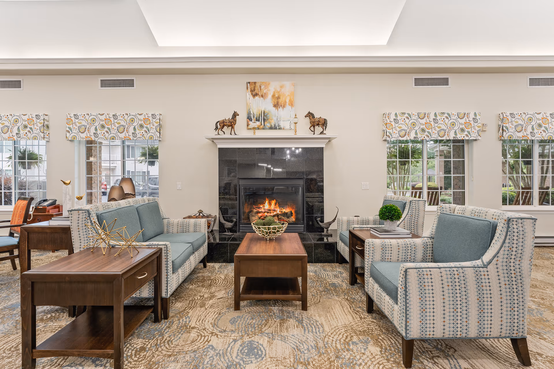 A cozy living room area with a lit fireplace in the center, flanked by two patterned armchairs and a matching sofa with blue cushions. A wooden coffee table and side tables with decorative items are placed in front of the seating. The room has large windows with patterned valances, allowing natural light to brighten the space.