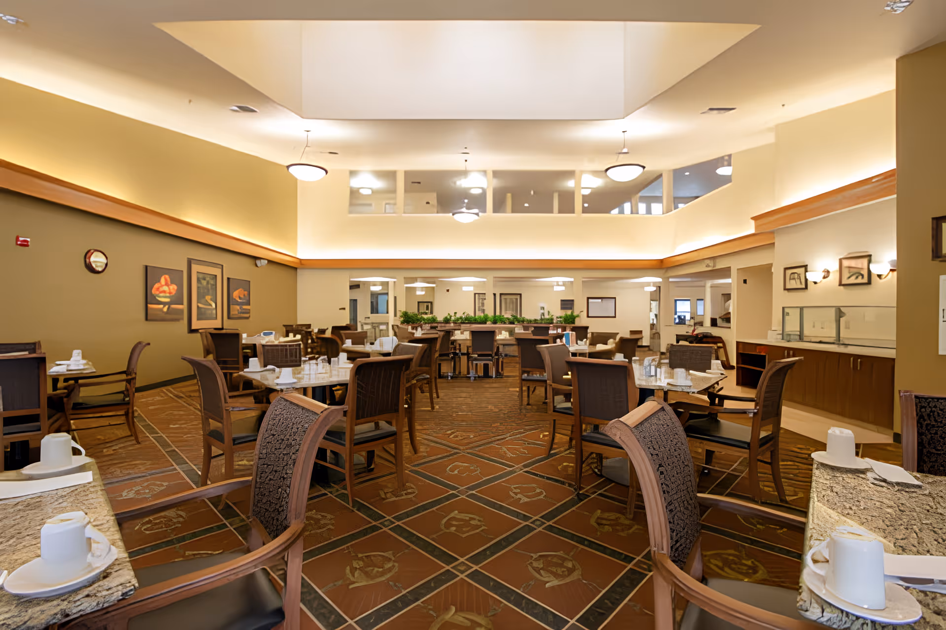 A spacious dining room in a senior living facility with multiple tables and chairs arranged neatly. The room features warm lighting, a high ceiling with a skylight, and decorative paintings on the walls. Tables are set with cups, napkins, and utensils, ready for meal service.