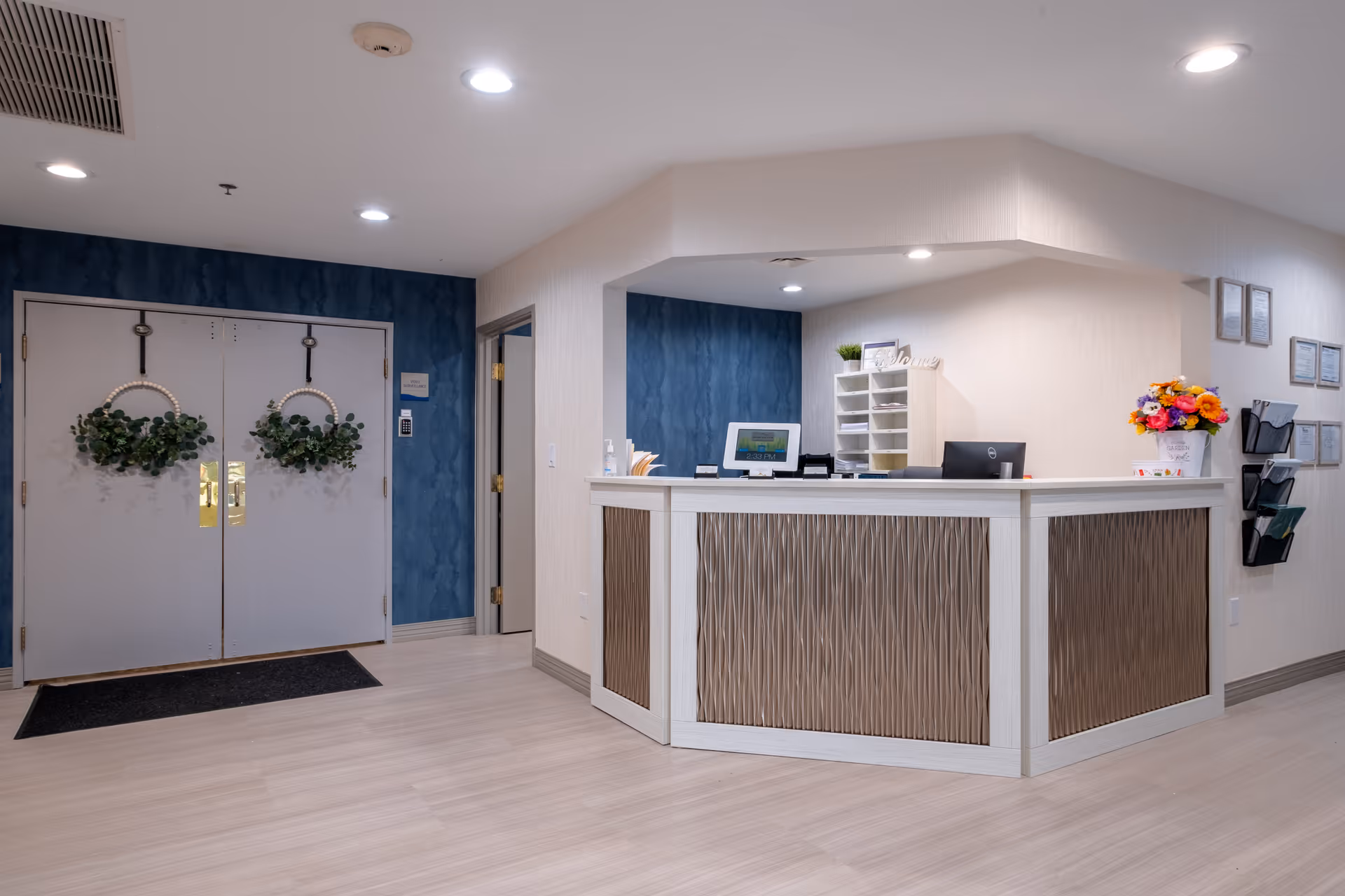 Reception area of Walnut Ridge Memory Care facility featuring a modern front desk with decorative paneling, a computer, and a flower arrangement. Behind the desk are shelves with mail slots and a small plant. To the left, double doors decorated with green wreaths hang on a blue accent wall. The floor is light wood, and the ceiling has recessed lighting.